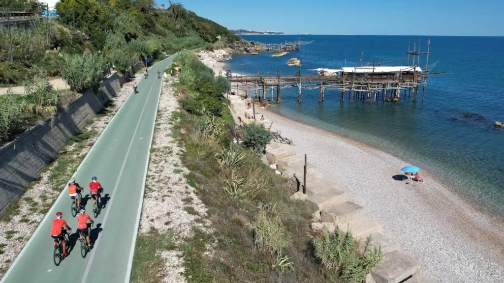 Cicloescursionisti del CAI pedalano sulla Via Verde della Costa dei Trabocchi con un trabocco sullo sfondo.